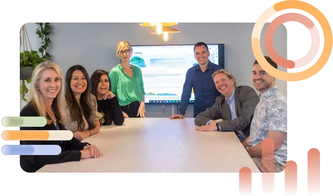 Group of people sitting and standing around a conference table, smiling toward the camera, with a screen and plants in the background, suggesting a collaborative office environment.