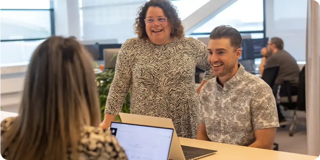 Three colleagues collaborating around a laptop in an office space, smiling and engaged.