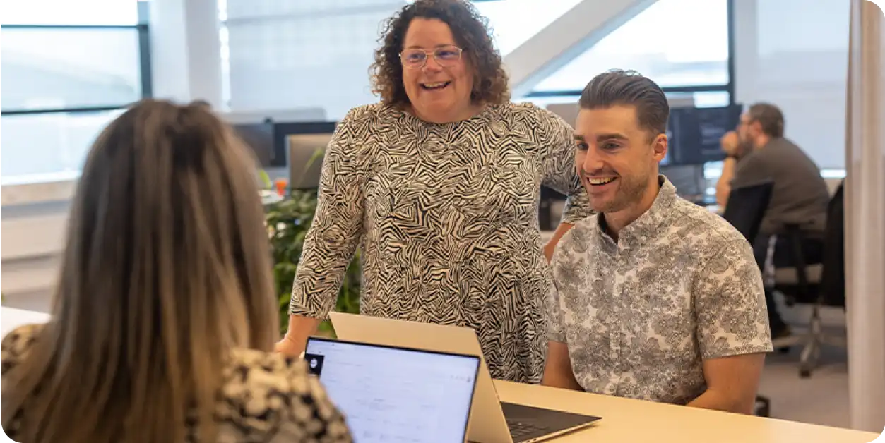 Three colleagues collaborating around a laptop in an office space, smiling and engaged.