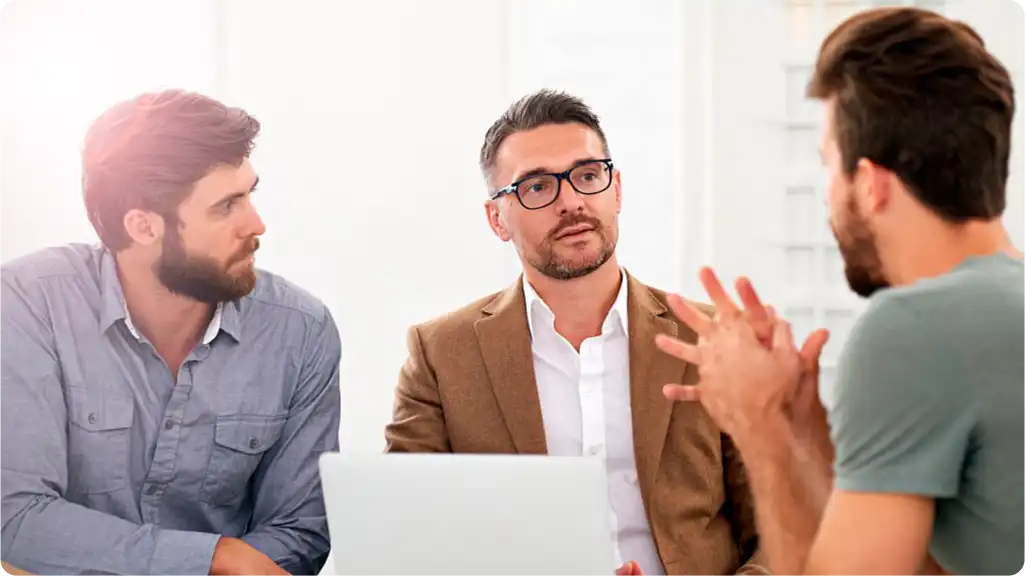 A business meeting or discussion between three men, with a laptop present on the table.