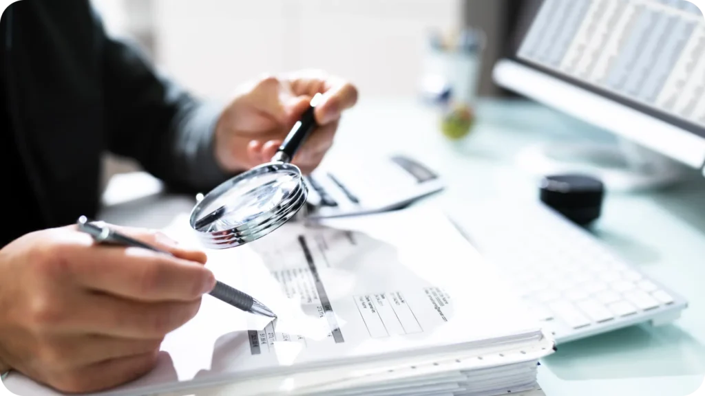 Close-up of a professional using a magnifying glass to review financial documents, symbolizing attention to detail and compliance management.