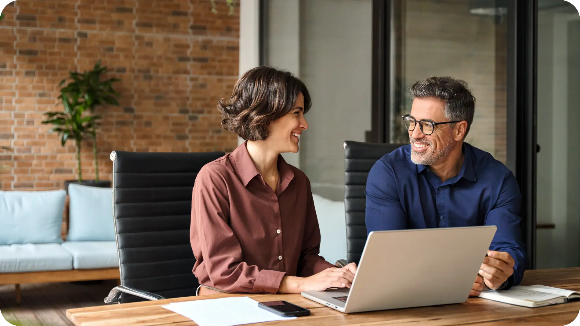 Two professionals collaborating in an office, smiling and discussing work while using a laptop, symbolizing teamwork and efficiency.