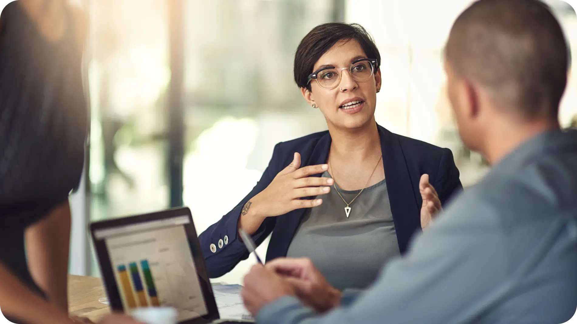 Business meeting scene with a confident woman in glasses speaking and gesturing, engaging in a discussion with two colleagues in a modern office setting. A laptop with a bar chart is visible on the table, suggesting a focus on analytics or performance metrics.