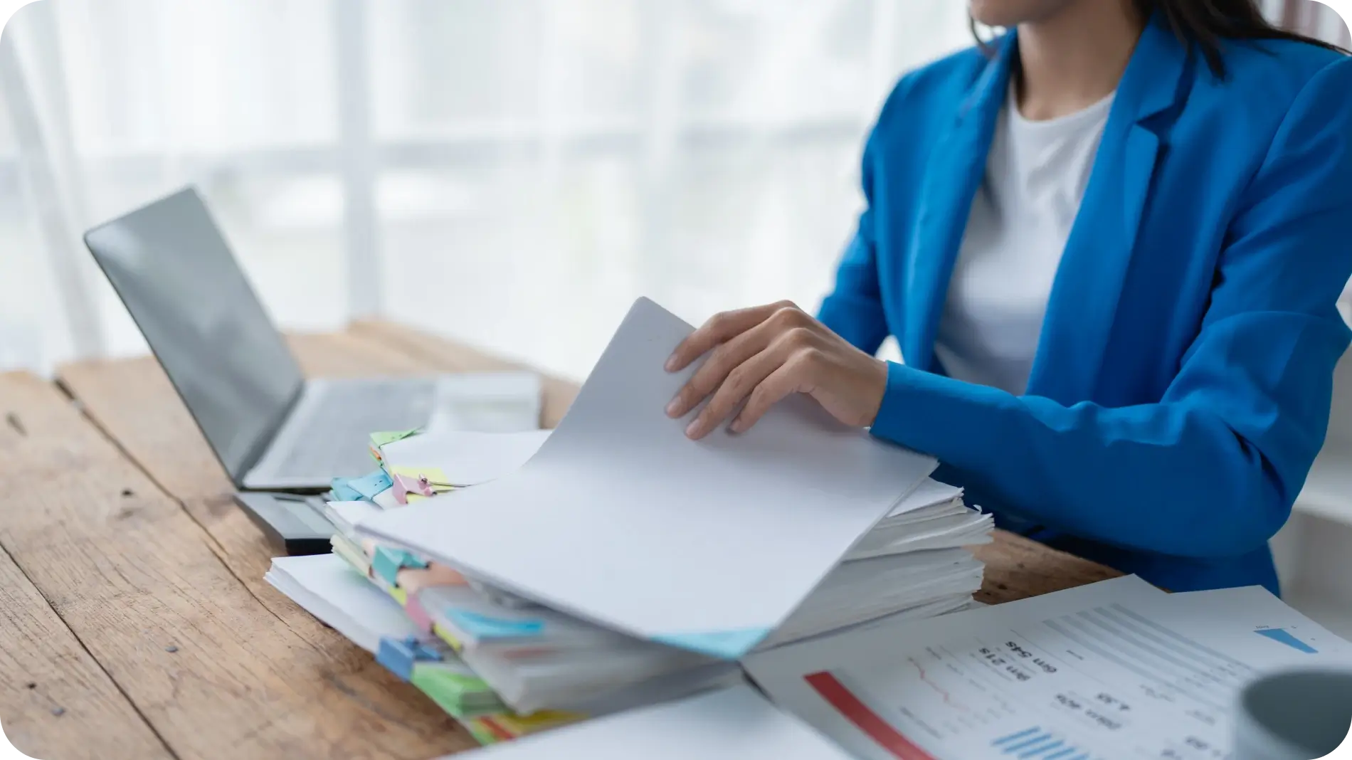 Woman sitting with a big pile of paperwork in front of a laptop.