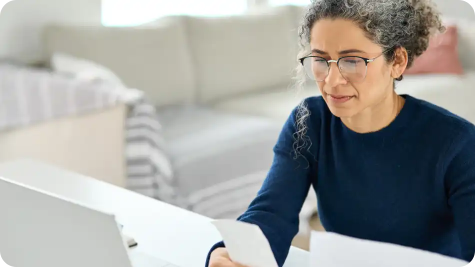 Person wearing glasses and a dark sweater sitting at a desk, looking at papers with a laptop open in front, in a cozy home environment.