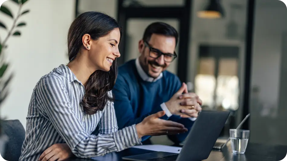 A smiling woman in a striped shirt discusses something on her laptop with a colleague in glasses and a sweater in a modern office setting.