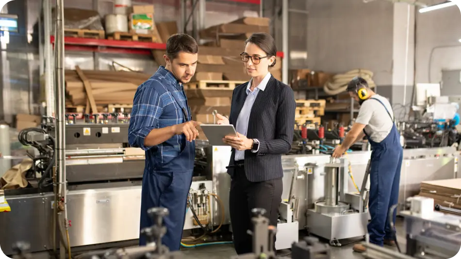 A factory setting where a manager with a tablet discusses work with a worker in overalls, while another operates machinery in the background.