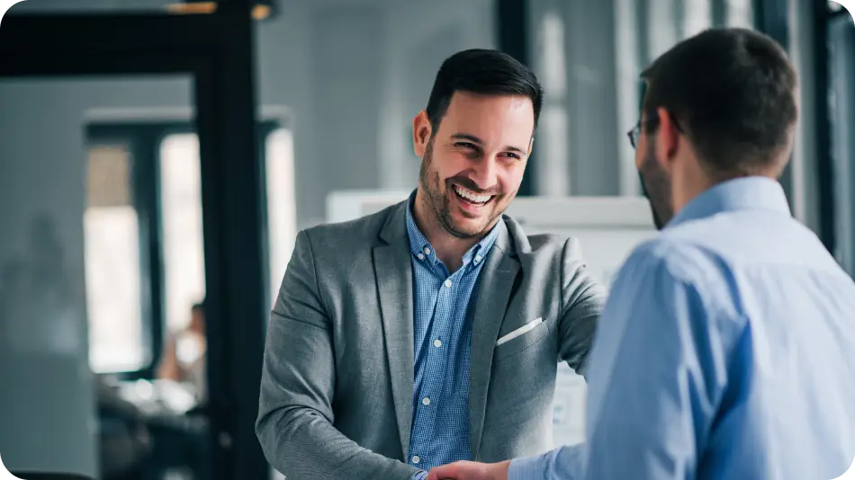 A smiling businessman in a gray blazer shakes hands with a colleague in an office setting, conveying a friendly and professional interaction.
