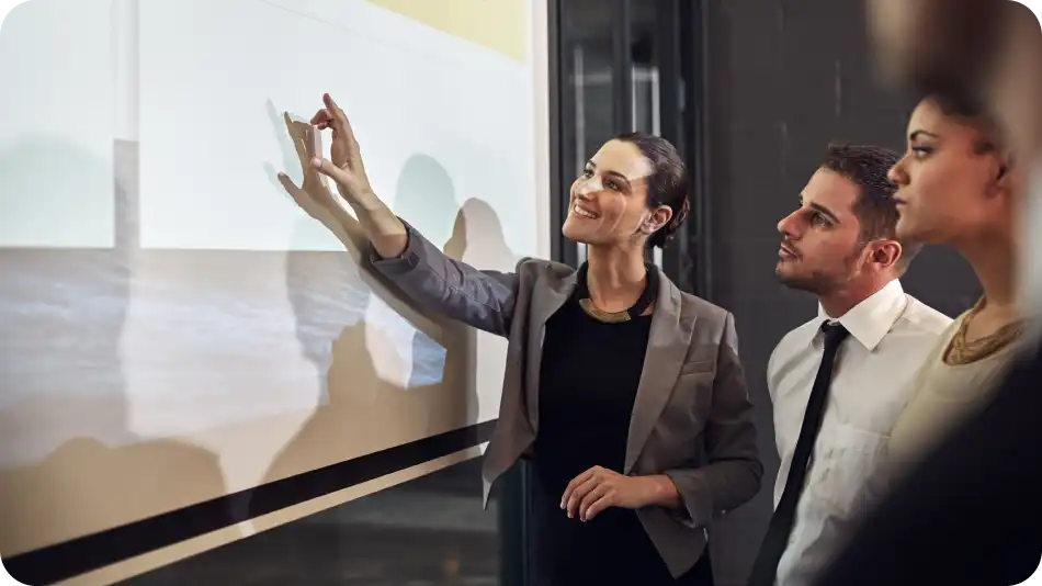 A woman in a blazer presents confidently, pointing at a screen while two colleagues listen attentively.