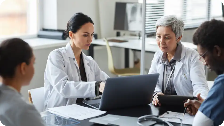 A group of medical professionals wearing lab coats are gathered around a table in a modern office. One doctor is working on a laptop while others review documents and take notes, engaged in a discussion.