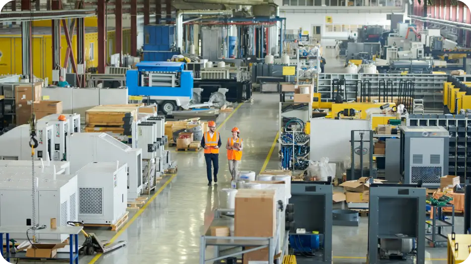 A large industrial manufacturing facility with various machines, equipment, and workstations. Two workers wearing safety vests and helmets walk along a clean, well-organized aisle, discussing production processes.