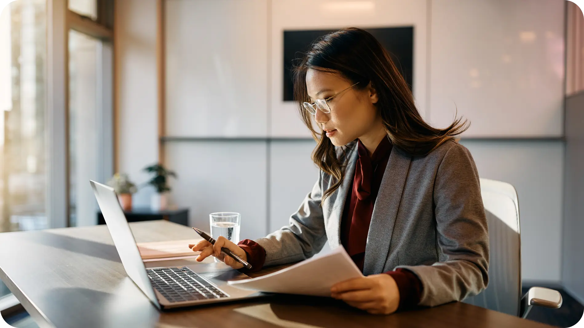 A professional woman in a gray blazer and glasses working at a desk in a modern office. She is focused on her laptop while holding a pen and reviewing documents, with a glass of water and office supplies nearby.