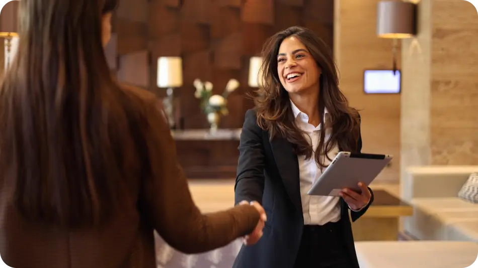 A smiling businesswoman in a black blazer shakes hands with another person while holding a tablet in a stylish office or hotel lobby.