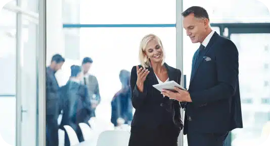 A professionally dressed man and woman in business suits are engaged in a conversation in a modern office setting. The woman is smiling while holding a phone, and the man is reviewing a document on a tablet. Other professionals are visible in the background.