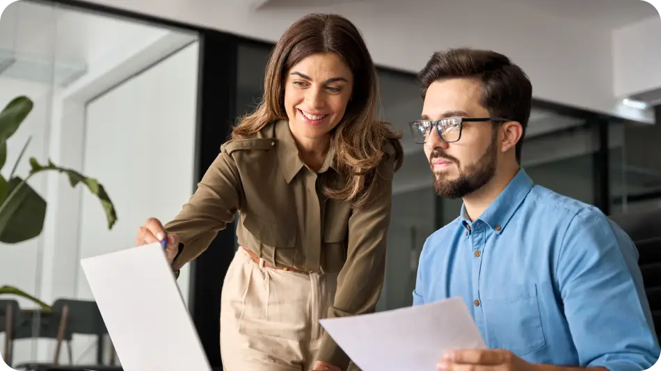 A professional woman in an olive-green blouse is smiling and pointing at a document while assisting a bearded man in glasses and a blue shirt. They are collaborating in a modern office setting with a sleek, minimalist design.