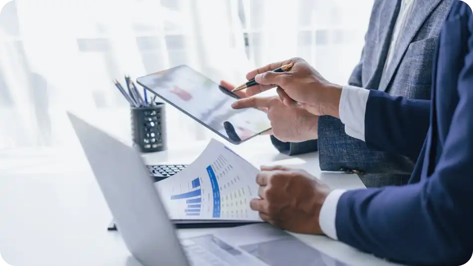 Close-up of two business professionals in suits analyzing financial documents, charts, and graphs. One person is holding a tablet while the other points with a pen, with a laptop and office supplies on the desk.