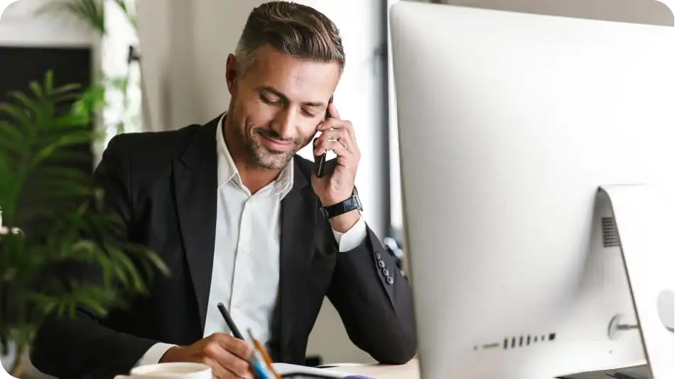 A professional businessman in a black suit and white shirt is sitting at his desk, talking on the phone while taking notes. He has a slight smile and is working in a modern office with a large computer monitor and greenery in the background.