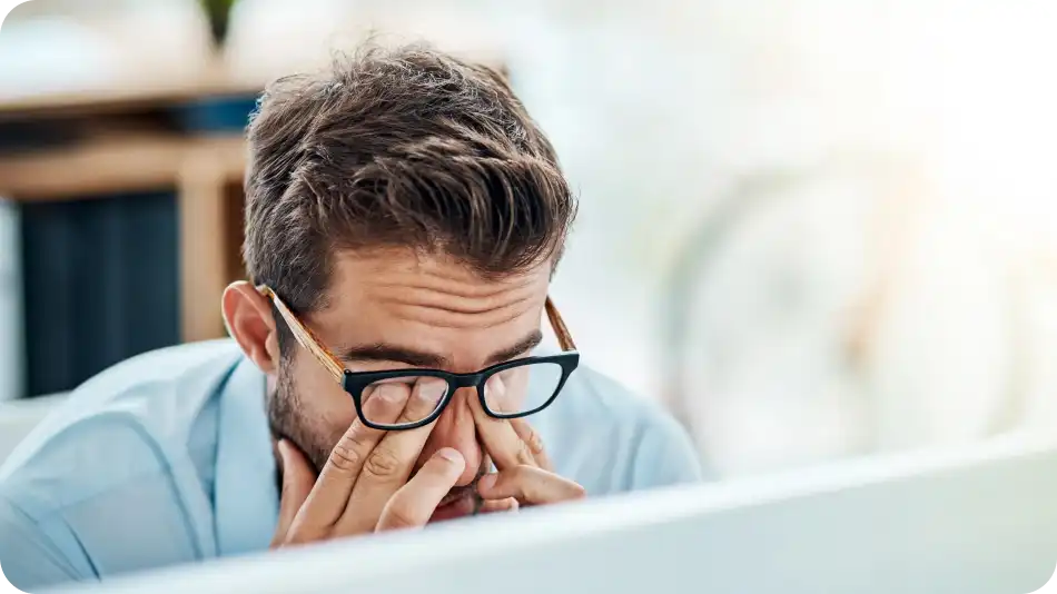 A stressed businessman wearing glasses and a light blue shirt sits at his desk, rubbing his eyes in frustration or fatigue. The blurred office background suggests a workplace setting with natural lighting.