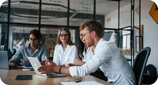 A group of young professionals in business casual attire are engaged in a discussion around a conference table in a modern office. One man in a white shirt is gesturing while presenting something on his laptop, while the others listen attentively.