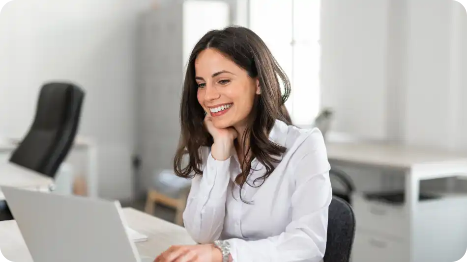 A smiling businesswoman with long dark hair, wearing a white blouse, is working on a laptop in a bright, modern office. She appears engaged and happy, with an open workspace and office furniture in the background.