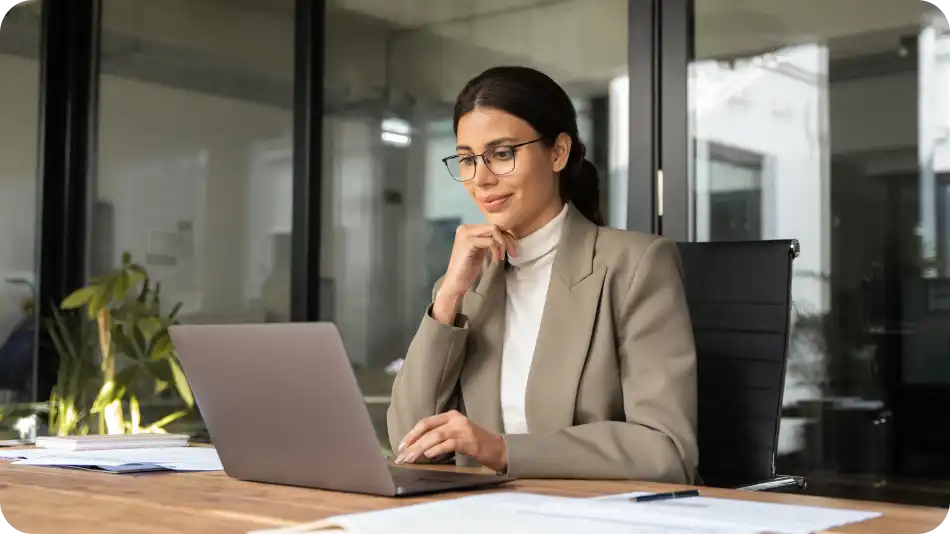 A professional woman wearing glasses and a beige blazer is working on a laptop in a modern office. She has a focused expression, with documents spread on the desk, and large windows in the background allowing natural light into the space.