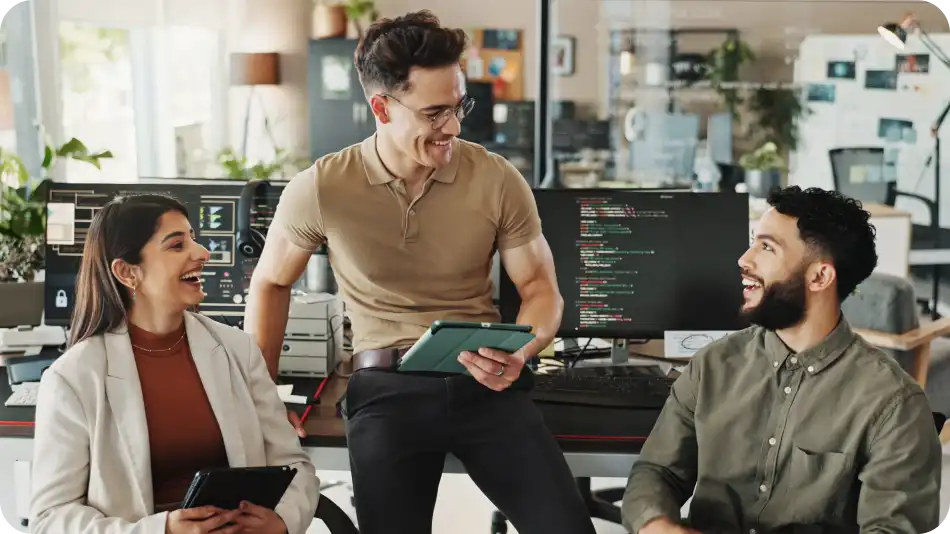 A group of three IT professionals in a modern office, engaged in a friendly discussion. A man in a brown polo shirt holds a tablet while talking to a seated male colleague with a beard and a female colleague in a white blazer. Computer screens displaying code are visible in the background.