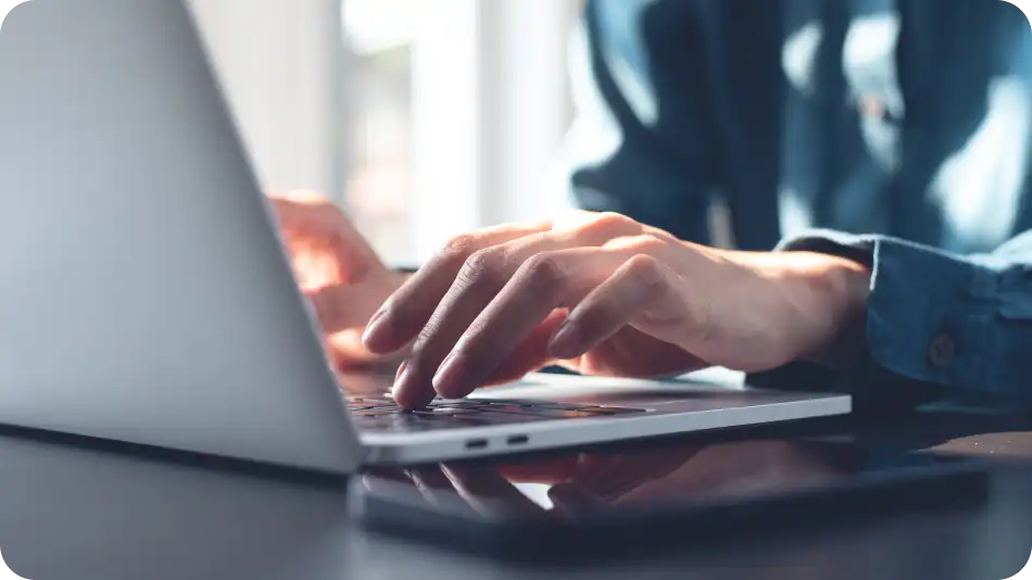 "Close-up of a person's hands typing on a laptop keyboard. The individual is wearing a dark blue shirt, and a smartphone is placed on the desk nearby, indicating a modern digital workspace.