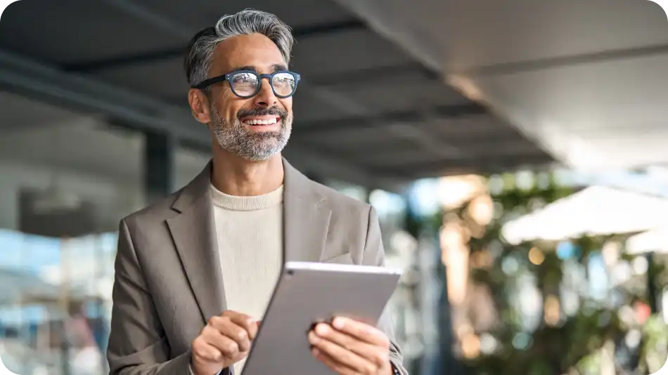 "A smiling middle-aged businessman with glasses and salt-and-pepper hair is holding a tablet outdoors. He is wearing a beige blazer and sweater, standing in a modern setting with greenery and glass architecture in the background.