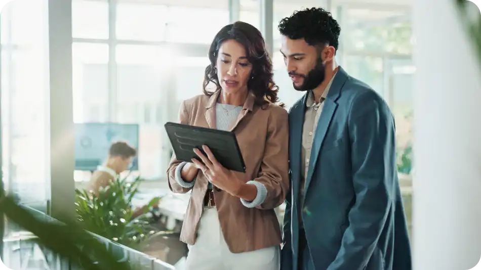 "A professional woman and man in business attire are standing in a modern office, reviewing information on a tablet. The woman is explaining something while the man listens attentively. Large windows, greenery, and an open workspace are visible in the background.