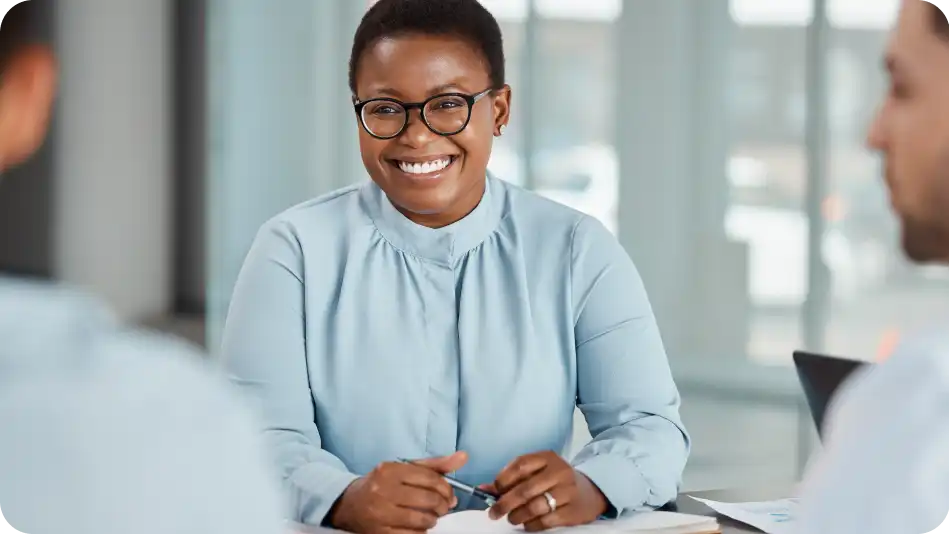 A smiling professional woman wearing glasses and a light blue blouse sits at a table during a business meeting. She is engaged in conversation with two colleagues, with documents and a laptop visible in a modern office setting.