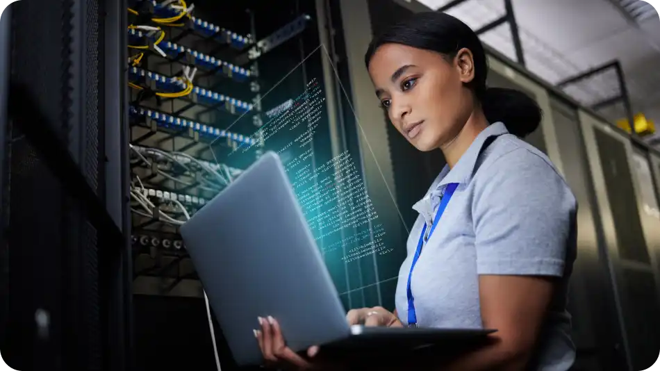A female IT professional in a gray polo shirt stands in a data center, working on a laptop. She is focused as she monitors network servers with cables and glowing indicators in the background, with a digital overlay of code displayed in front of her.