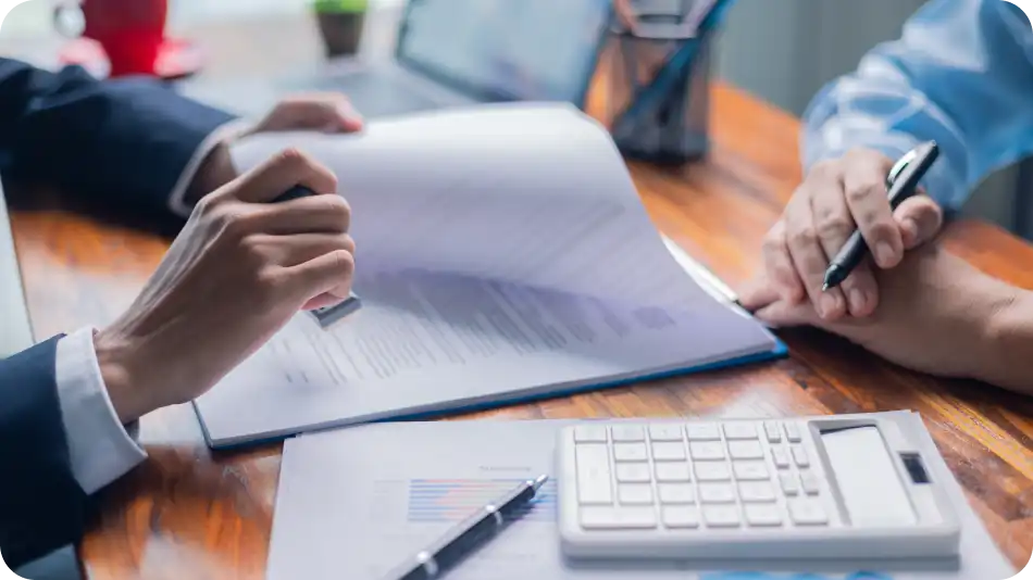 Close-up of two professionals in business attire reviewing documents at a wooden desk. One person is holding a stamp, while the other is holding a pen. A calculator, financial charts, and a laptop are also present on the desk.