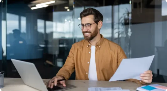 A smiling man with glasses and a beard, wearing a brown shirt, sitting at a modern office desk. He is holding a paper in one hand while using a laptop with the other. The office has glass partitions and a contemporary design with natural lighting.