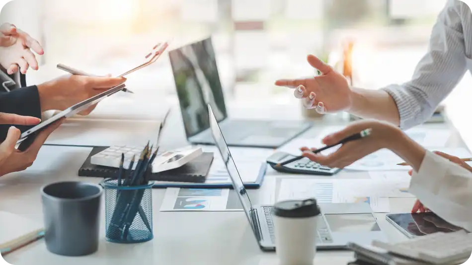 A close-up of a business meeting where multiple professionals are discussing financial documents, using laptops, tablets, and calculators. Hands are gesturing and holding pens, indicating an active discussion. Office supplies like coffee cups and pen holders are on the table.