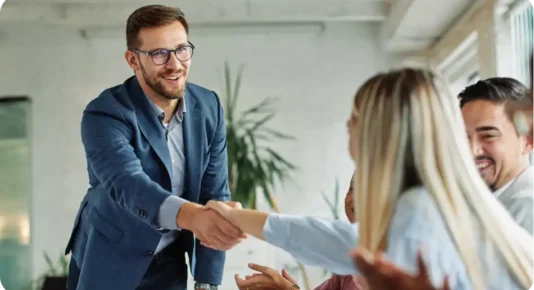 A smiling businessman in a blue blazer and glasses is shaking hands with a blonde woman in a light blue shirt during a meeting in a modern office. Other colleagues are seated nearby, smiling and engaged in the conversation.