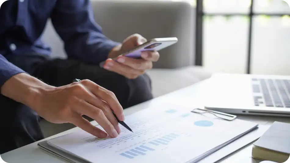 A close-up of a businessman wearing a dark shirt, holding a smartphone in one hand and a pen in the other while analyzing financial documents with charts and graphs. A laptop, notebook, and paperclip are on the desk in a well-lit office setting.