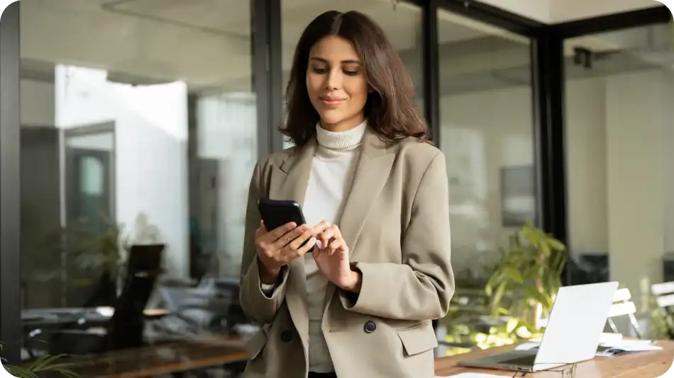 A professional woman in a beige blazer and white turtleneck is standing in a modern office, looking at her smartphone with a slight smile. A laptop and office decor, including plants, are visible in the background.