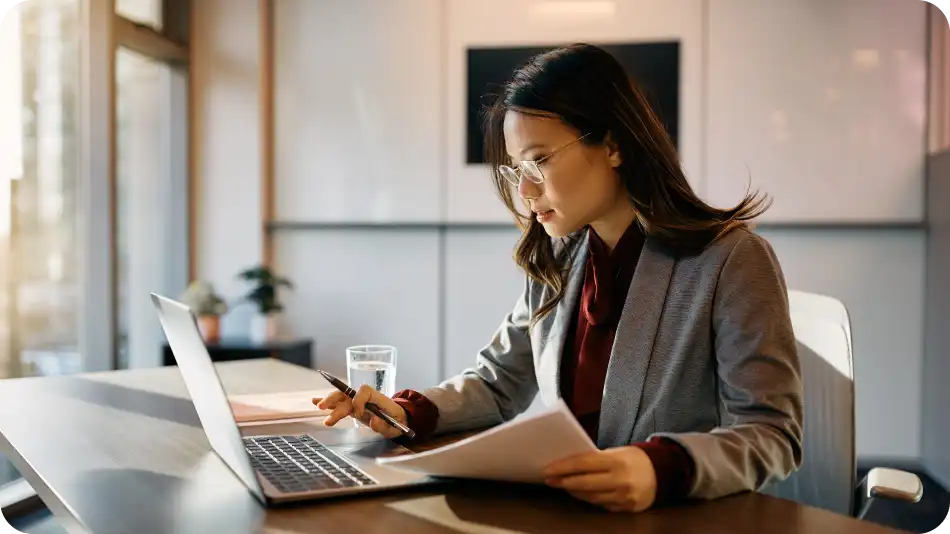 A professional woman wearing glasses and a gray blazer is sitting at a desk in a modern office. She is focused on her laptop while holding a pen and reviewing paperwork. A glass of water and office decor are visible in the background.