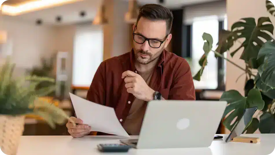 Man with glasses reviewing a document while working on a laptop at a modern desk.