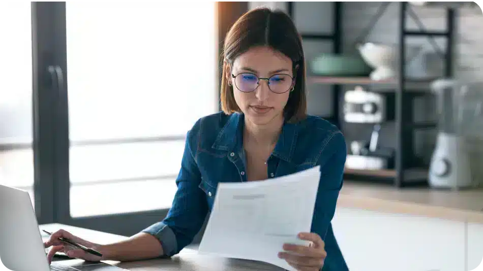 Woman wearing glasses reviewing a document while working on a laptop at home.