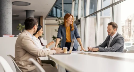 A group of colleagues are engaged in a meeting, with one woman standing and speaking while others listen and take notes.