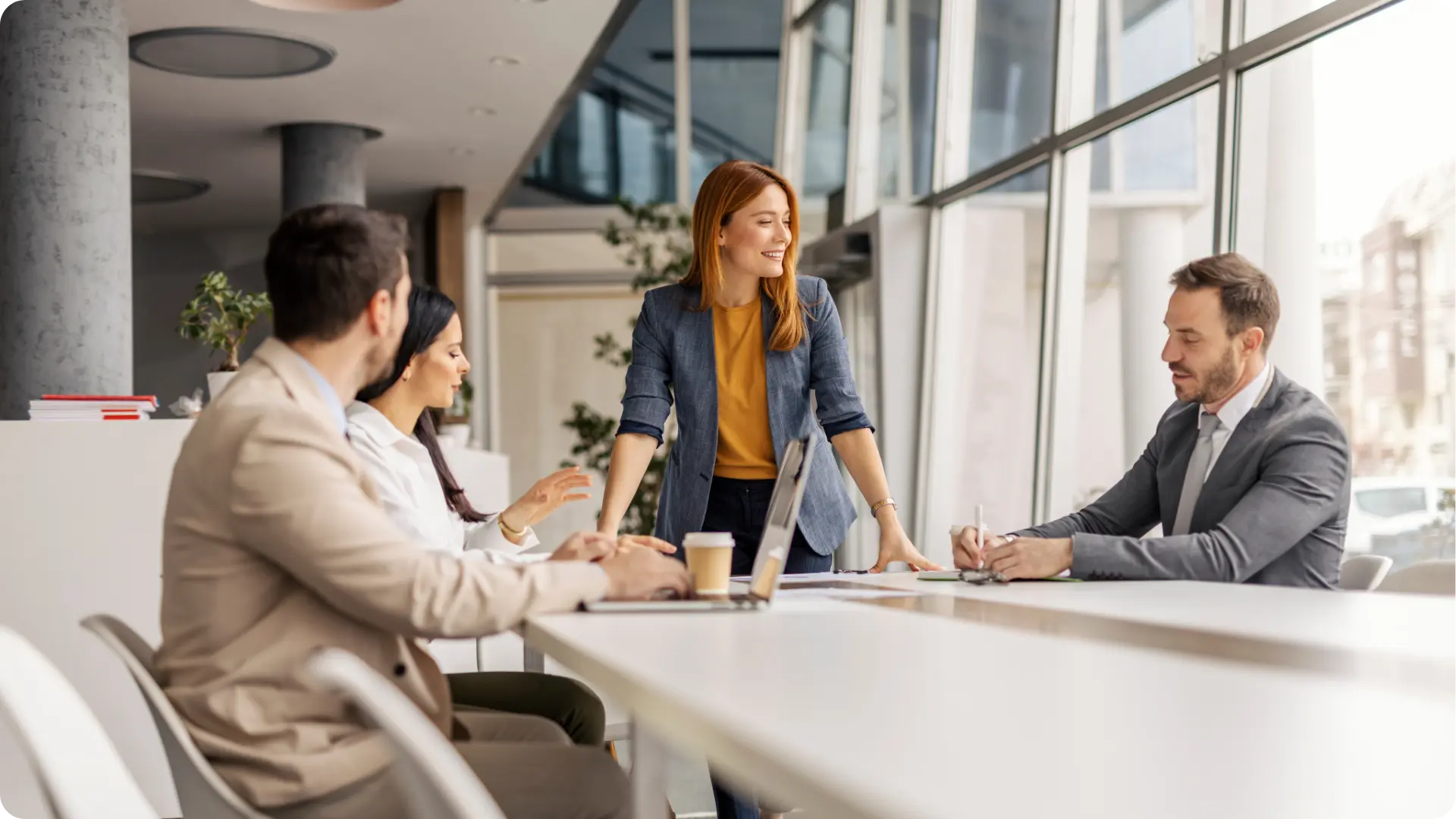 A group of colleagues are engaged in a meeting, with one woman standing and speaking while others listen and take notes.