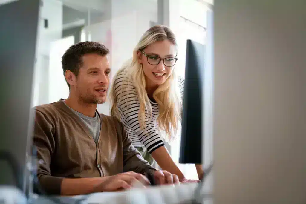 A man and woman are smiling while looking at a computer screen, suggesting collaboration or shared focus on a task.